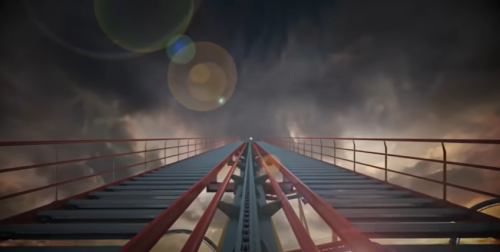 View looking up a roller coaster track towards a dramatic sky with clouds and lens flares.