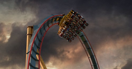 A roller coaster reaches the top of a loop, with riders experiencing excitement against a dramatic sky.