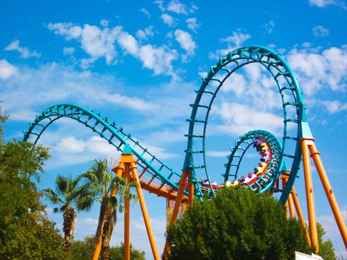 A colorful roller coaster twists against a blue sky with fluffy clouds, surrounded by palm trees and greenery.
