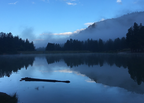 A serene lake surrounded by misty mountains and pine trees, reflecting the blue sky and clouds at dawn.