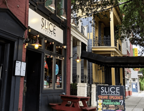 Exterior of Slice Pizzeria with a red picnic table and a chalkboard menu outside, surrounded by colorful buildings.