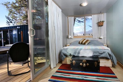 Cozy bedroom with a bed, colorful rug, and large windows overlooking nature, featuring a vintage trunk and a chair.