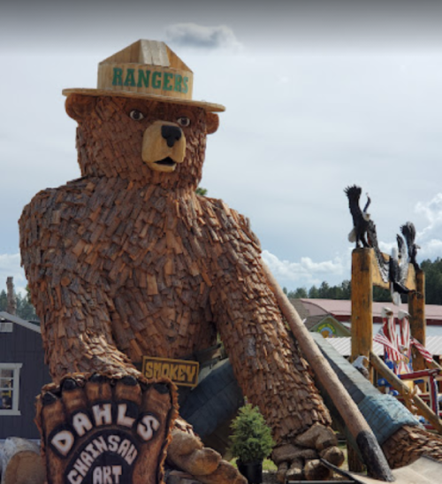 A large wooden bear sculpture wearing a ranger hat, surrounded by other wooden art pieces in an outdoor setting.