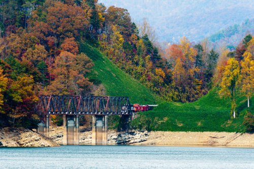 A red train crosses a metal bridge over a calm lake, surrounded by colorful autumn foliage and rolling hills.