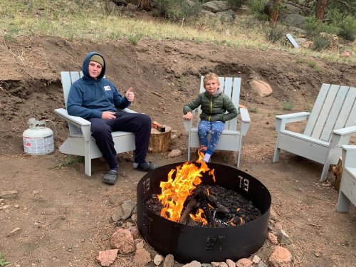 Two people sit by a campfire in outdoor chairs, enjoying the warmth and scenery in a natural setting.