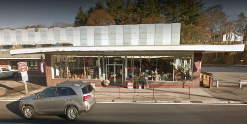 A storefront with large windows, displaying various items, and a gray SUV parked in front on a sunny day.