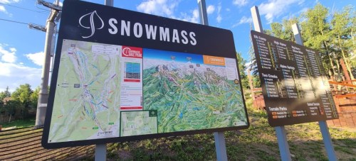 Map of Snowmass, displaying trails, lifts, and terrain features, with a scenic backdrop of trees and mountains.