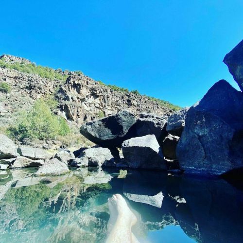 A serene natural hot spring surrounded by rocks and greenery, with a person's legs visible in the water.