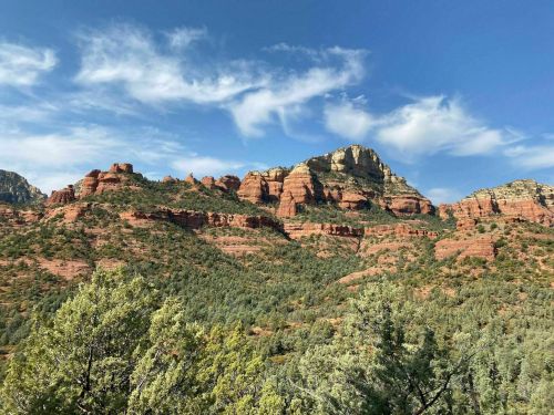 Red rock formations rise against a blue sky, surrounded by green trees in a scenic landscape.