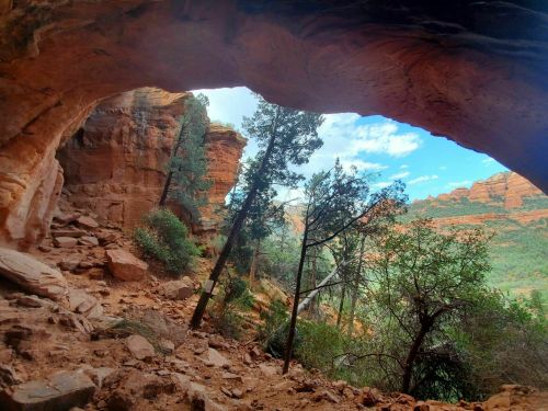 View through a natural rock arch, showcasing a scenic landscape of trees and red rock formations under a blue sky.