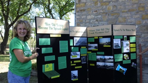 A woman stands beside a display board titled "How Did Larned Become The Spinach Capital?" with photos and text.