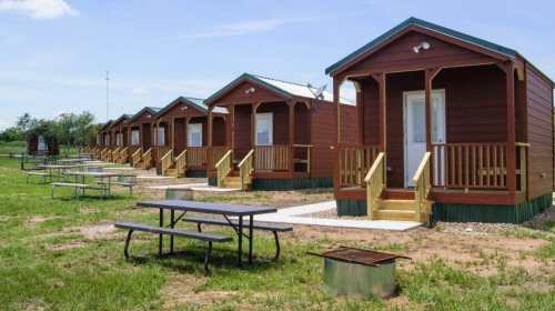 Row of rustic cabins with porches, surrounded by green grass and picnic tables under a clear blue sky.
