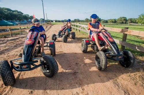 Three children riding go-karts on a dirt track, surrounded by greenery and wooden fencing.