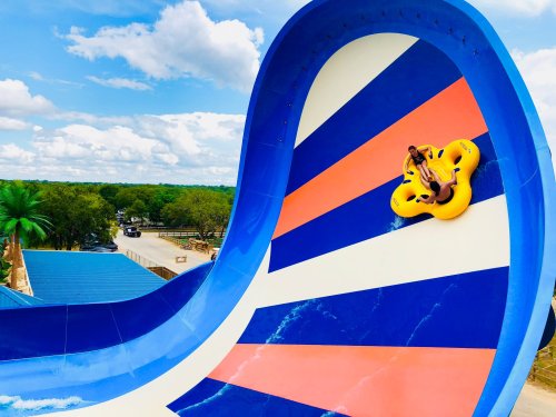 A person on a yellow inner tube slides down a colorful water slide against a blue sky and green landscape.