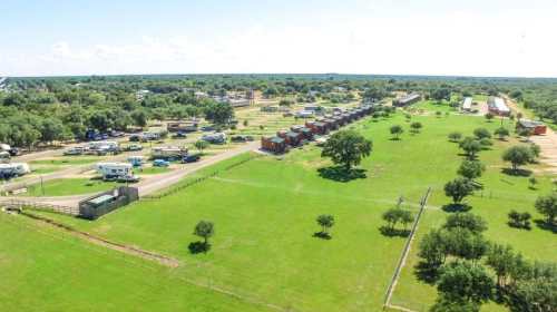 Aerial view of a rural area with green fields, scattered trees, and rows of buildings or trailers along a road.