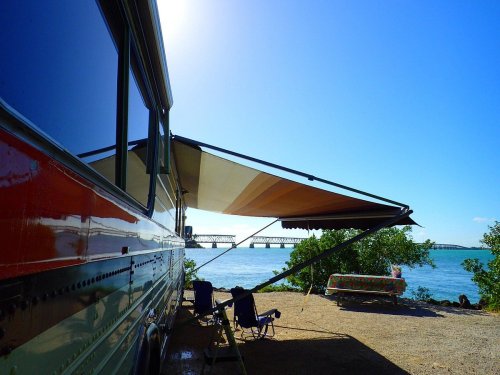A colorful RV parked by the water, with an awning extended and chairs set up, overlooking a bridge in the distance.