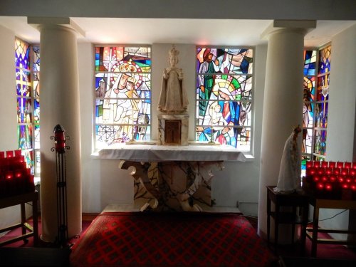 A serene chapel interior featuring stained glass windows, a statue on an altar, and red candles on either side.