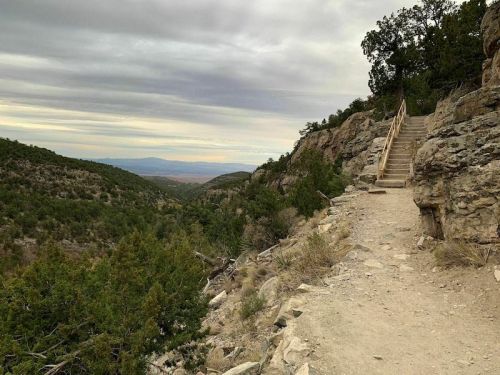 A rocky trail leads up a hillside with wooden steps, surrounded by greenery and distant mountains under a cloudy sky.