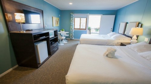 A cozy hotel room with two beds, a TV unit, and a small table by the window, featuring blue walls and natural light.