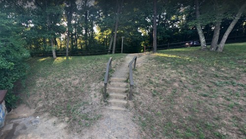A grassy area with a set of stone steps leading up to a wooded area, surrounded by trees and a fence.