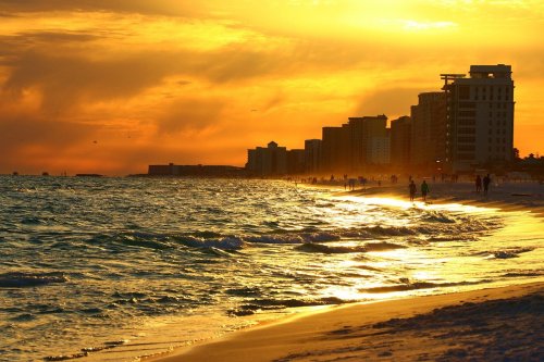 Sunset over a beach with golden waves, silhouetted buildings, and people walking along the shore.