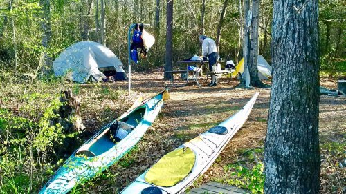 Two kayaks are parked near a campsite with tents and a picnic table surrounded by trees.