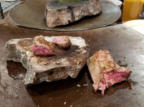 Two pieces of cooked meat resting on a hot stone surface, with a pink, juicy interior visible.