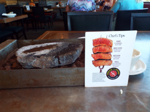 A stone cooking platter with a menu card displaying chef's tips for cooking meat, set on a restaurant table.