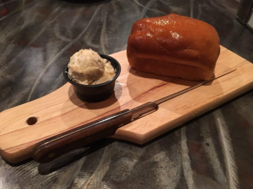 A wooden board with a loaf of bread and a small bowl of butter, accompanied by a knife.