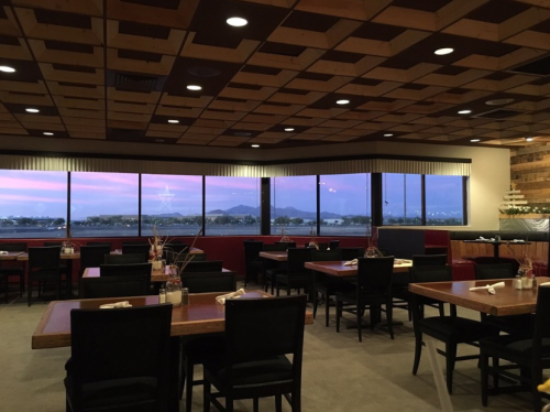 Interior of a restaurant with wooden tables, black chairs, and large windows showcasing a scenic view.