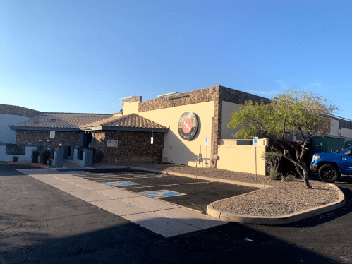 A stone-clad building with a logo, surrounded by a parking lot and desert landscaping under a clear blue sky.