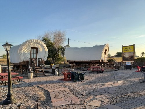 Two covered wagons on a gravel path, surrounded by picnic tables and a sign for a campground in a sunny outdoor setting.