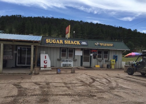 A rustic building with a sign reading "Sugar Shack," offering breakfast and burgers, surrounded by trees and dirt.