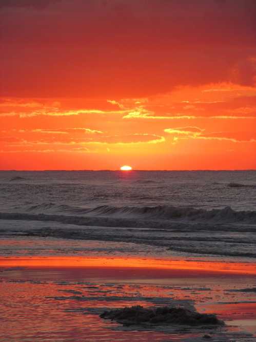 A vibrant sunset over the ocean, with orange and red hues reflecting on the water and gentle waves lapping at the shore.