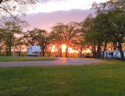 A serene campsite at sunset, with RVs parked among trees and a colorful sky in the background.