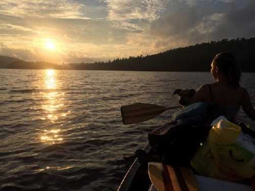 A person sits in a canoe at sunset, holding a dog, with calm water and trees in the background.