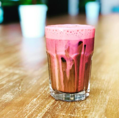 A glass of vibrant pink drink with frothy top, sitting on a wooden table. Blurred background with light colors.