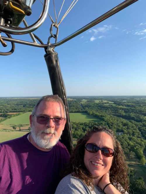 A couple smiles for a selfie in a hot air balloon, overlooking a lush green landscape below.