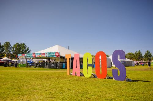 Colorful "TACOS" sign in front of a food festival tent on a sunny day with green grass.