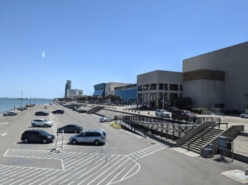 A sunny waterfront scene featuring a nearly empty parking lot and modern buildings along the shoreline.