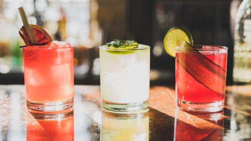 Three colorful cocktails on a bar: a red drink with a lime, a green drink with a cucumber, and a pink drink with a garnish.