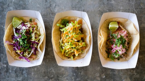 Three tacos in white trays, each topped with colorful ingredients like cabbage, cheese, and cilantro, with lime wedges.