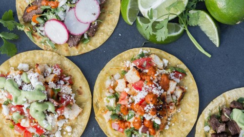 A variety of colorful tacos topped with radishes, cilantro, lime, and cheese on a dark surface.
