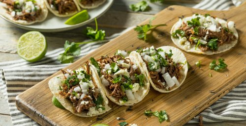 A wooden board with three tacos filled with shredded meat, onions, and cilantro, garnished with lime wedges.