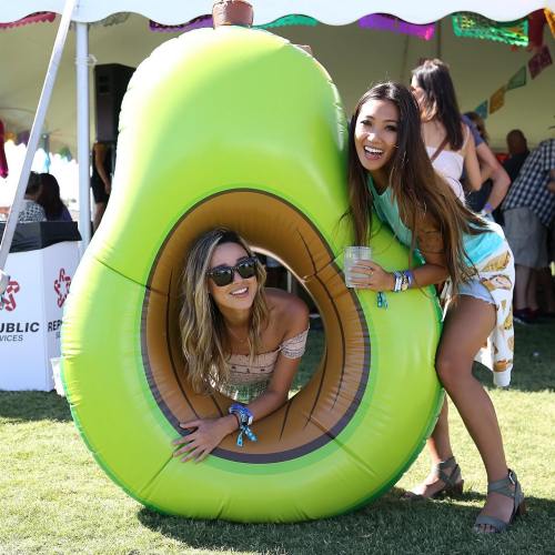 Two women pose playfully inside a large inflatable avocado at an outdoor event, smiling and holding drinks.