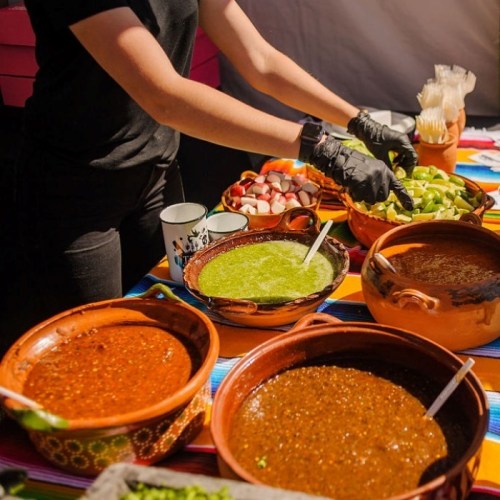 A person in gloves prepares various colorful salsas and toppings on a vibrant tablecloth at a food event.