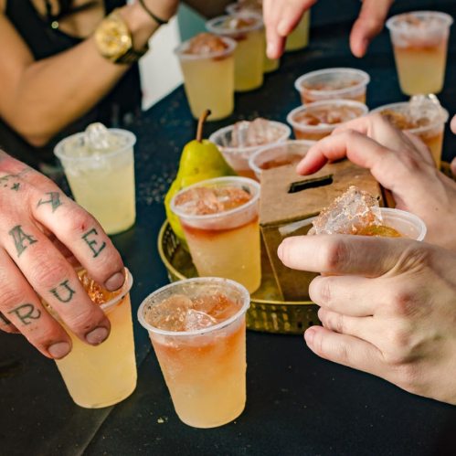 A table with various drinks in plastic cups, hands reaching for cups, and a pear on the side.