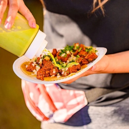 A person holds a plate of tacos topped with meat, onions, and cilantro, drizzling green sauce on top.