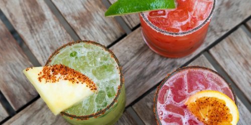Three colorful cocktails on a wooden table, garnished with fruit and rimmed with salt.