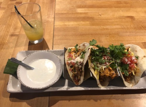 Three tacos topped with cilantro and cheese, served with a side of sauce and a drink on a wooden table.